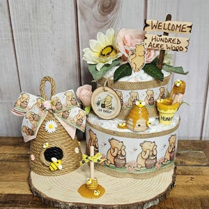 May include: A Winnie the Pooh themed diaper cake with a honey pot, bees, and a sign that says "Welcome to the Hundred Acre Wood".