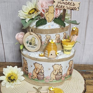 May include: A two-tiered diaper cake decorated with Winnie the Pooh and honey pot themes. The cake is adorned with flowers, a wooden sign that reads "Welcome to the Hundred Acre Wood", and a wooden honey dipper.