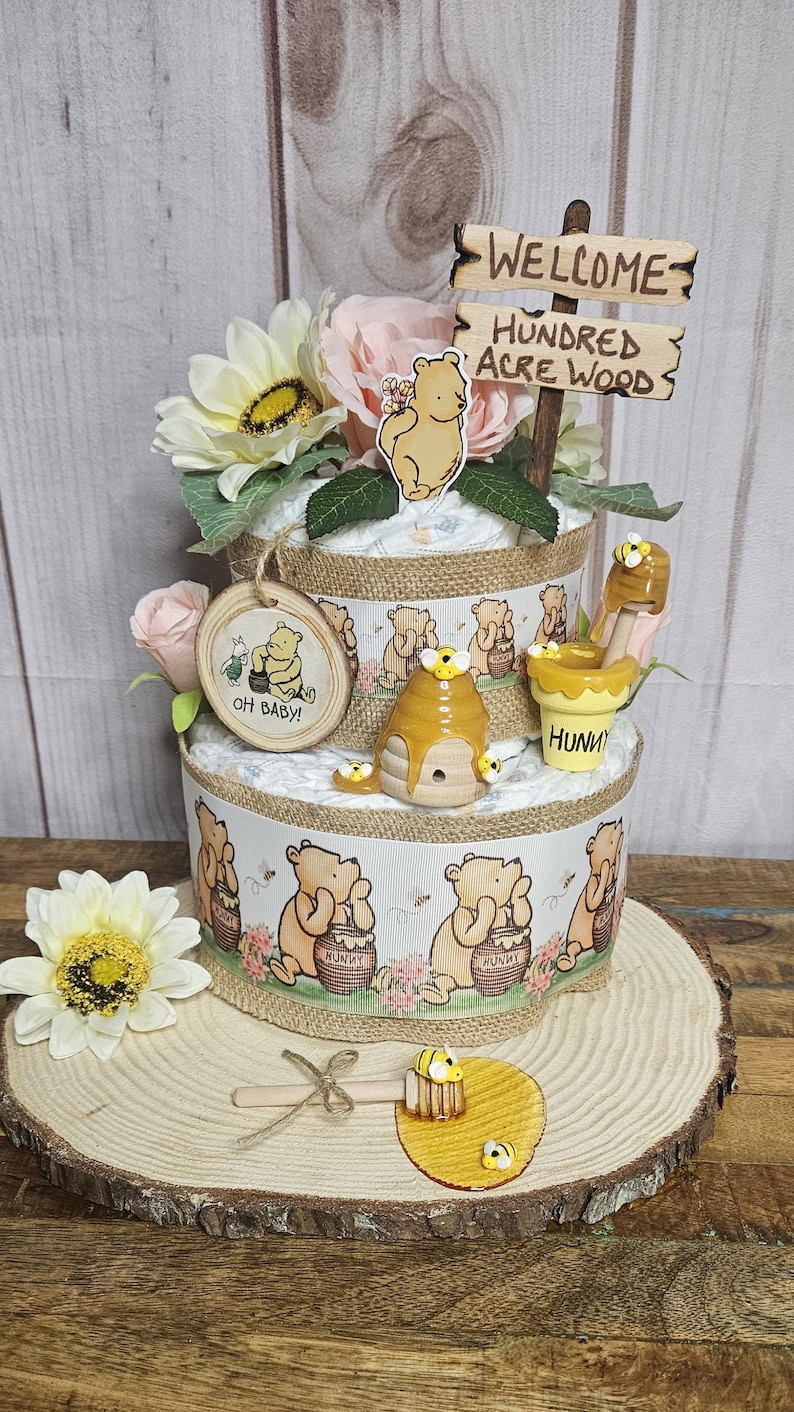 May include: A two-tiered diaper cake decorated with Winnie the Pooh and honey pot themes. The cake is adorned with flowers, a wooden sign that reads "Welcome to the Hundred Acre Wood", and a wooden honey dipper.