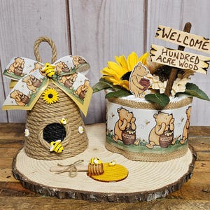 May include: A Winnie the Pooh themed baby shower centerpiece with a honey pot, a beehive, and a diaper cake decorated with Winnie the Pooh characters. The diaper cake is wrapped in burlap and has a sunflower on top. The sign says "Welcome Hundred Acre Wood".