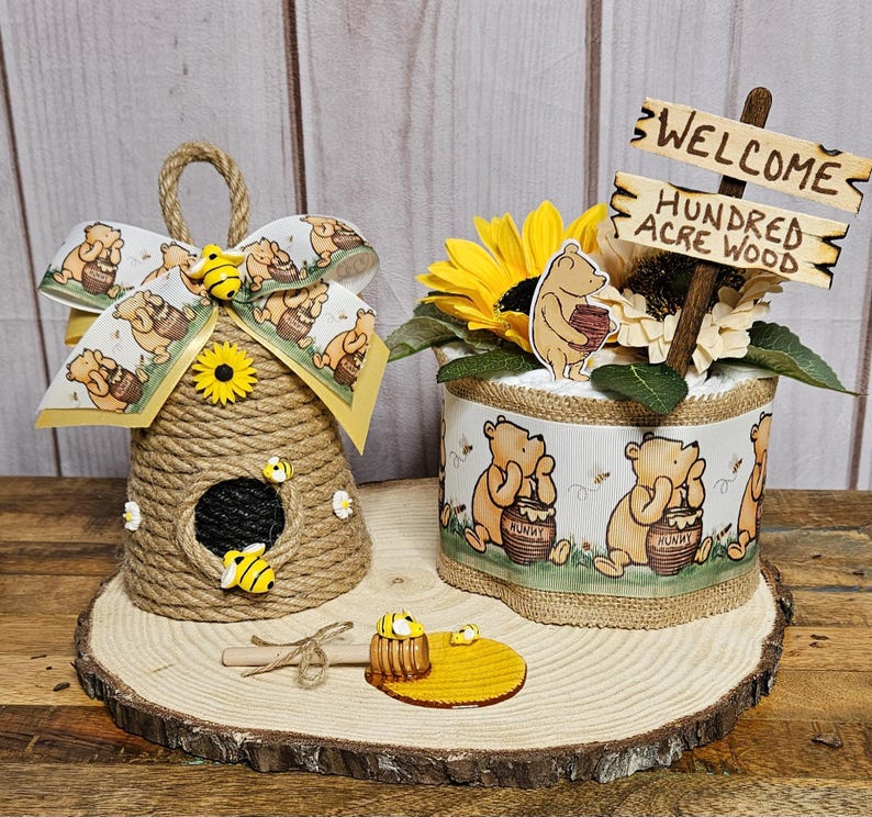May include: A Winnie the Pooh themed baby shower centerpiece with a honey pot, a beehive, and a diaper cake decorated with Winnie the Pooh characters. The diaper cake is wrapped in burlap and has a sunflower on top. The sign says "Welcome Hundred Acre Wood".
