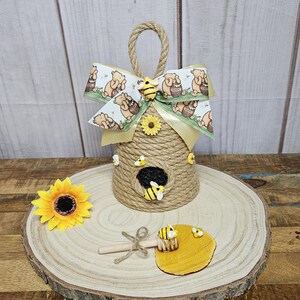 May include: A Winnie the Pooh themed beehive door hanger made of twine with a yellow ribbon bow and a honey dipper with a small pool of honey.