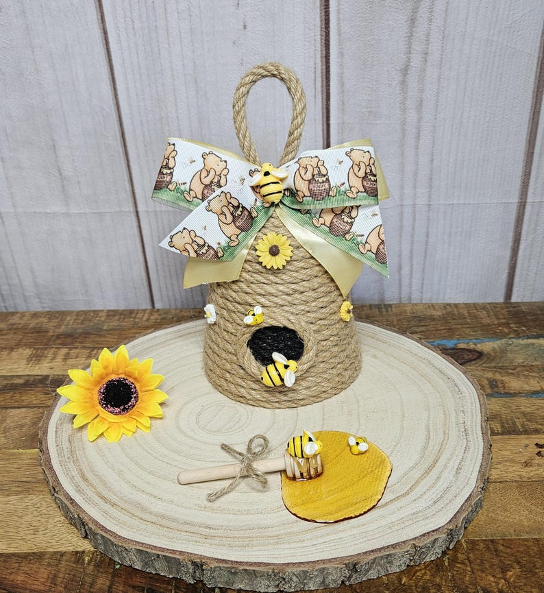 May include: A Winnie the Pooh themed beehive door hanger made of twine with a yellow ribbon bow and a honey dipper with a small pool of honey.