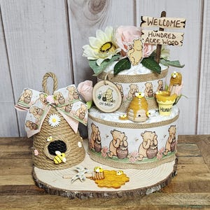 May include: A Winnie the Pooh themed diaper cake with a wooden sign that says "Welcome to the Hundred Acre Wood". The cake is decorated with Winnie the Pooh characters, honeycombs, and flowers. There is also a small beehive-shaped decoration with a bee on it.