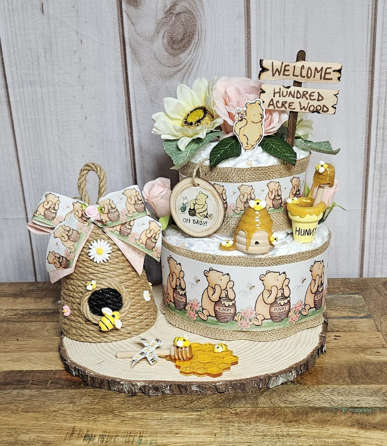 May include: A Winnie the Pooh themed diaper cake with a wooden sign that says "Welcome to the Hundred Acre Wood". The cake is decorated with Winnie the Pooh characters, honeycombs, and flowers. There is also a small beehive-shaped decoration with a bee on it.