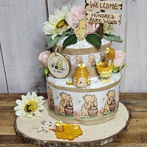 May include: A two-tiered Winnie the Pooh themed diaper cake decorated with Winnie the Pooh figurines, honeycombs, and flowers. The cake is topped with a wooden sign that reads "Welcome to the Hundred Acre Wood".