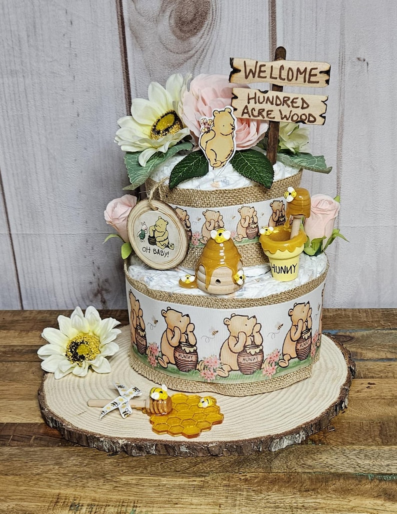 May include: A two-tiered Winnie the Pooh themed diaper cake decorated with Winnie the Pooh figurines, honeycombs, and flowers. The cake is topped with a wooden sign that reads "Welcome to the Hundred Acre Wood".