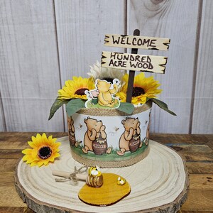 May include: A Winnie the Pooh themed baby shower centerpiece with sunflowers, a Winnie the Pooh figurine, and a sign that reads "Welcome to the Hundred Acre Wood".