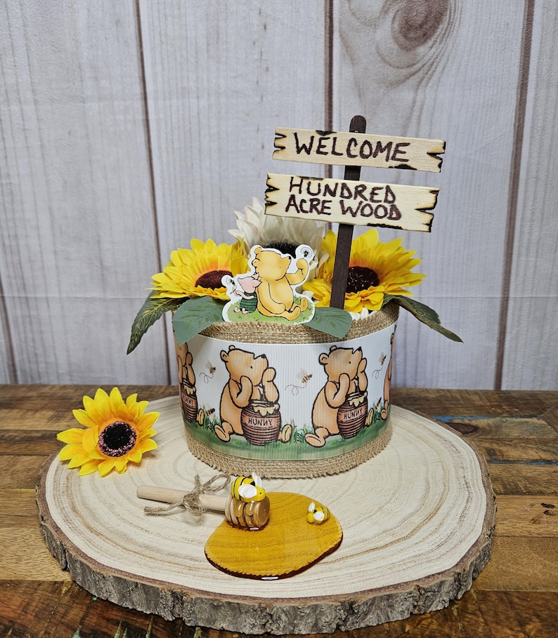 May include: A Winnie the Pooh themed baby shower centerpiece with sunflowers, a Winnie the Pooh figurine, and a sign that reads "Welcome to the Hundred Acre Wood".