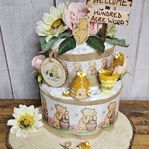 May include: A two-tiered Winnie the Pooh diaper cake decorated with yellow and pink flowers, a wooden sign that says "Welcome to the Hundred Acre Wood", and Winnie the Pooh figurines. The cake is decorated with a Winnie the Pooh print and a honey pot.