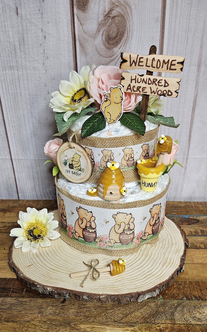 May include: A two-tiered Winnie the Pooh diaper cake decorated with yellow and pink flowers, a wooden sign that says "Welcome to the Hundred Acre Wood", and Winnie the Pooh figurines. The cake is decorated with a Winnie the Pooh print and a honey pot.
