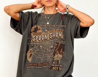 May include: A dark gray t-shirt with a vintage-style graphic print of a spooky Halloween show. The graphic features a skull and crossbones, skeletons dancing, and a Frankenstein's monster. The text reads "Salem Theater Presents Midnight Live on Stage Spook Show See Frankenstein's Monster In Person! Dancing Skeletons Witches & Real Ghosts One Night Only Oct 31."