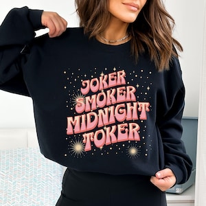 May include: Black sweatshirt with the words "Joker, Smoker, Midnight, Toker" in pink and peach retro font. The design includes starburst and star accents. The sweatshirt is a casual style.