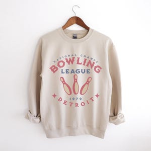 May include: Beige sweatshirt with the text "NATIONAL CHAMPS BOWLING LEAGUE DETROIT 1979" in red and blue. The design includes three bowling pins. The sweatshirt is hanging on a wooden hanger.