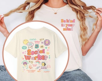 May include: A cream colored t-shirt with a graphic print of the words "Social Worker" surrounded by colorful cartoon images and text. The text "Be kind to your mind" is printed on the front of the shirt.
