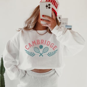 May include: White sweatshirt with a pink and blue graphic of two tennis rackets and a tennis ball with the word "CAMBRIDGE" in pink letters.