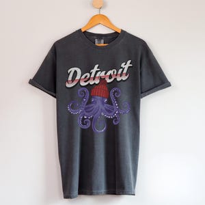 May include: A dark gray t-shirt with a graphic print of an octopus wearing a red and white striped beanie. The text "Detroit" is printed in white above the octopus.