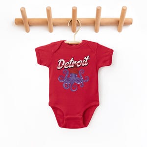 May include: Red baby onesie with a purple octopus wearing a red and white striped hat. The onesie has the word "Detroit" printed in a retro font.