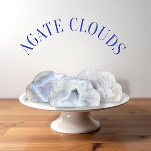 May include: Three cloud-shaped agate stones with blue and white patterns are displayed on a white pedestal cake stand. The text "AGATE CLOUDS" is arched above the stones in blue.