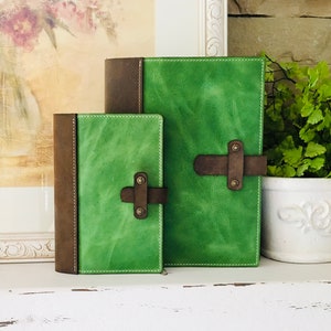 May include: Two leather-bound journals with green and brown covers. The larger journal has a brown leather strap with a brass buckle. The smaller journal has a similar strap but is smaller in size.