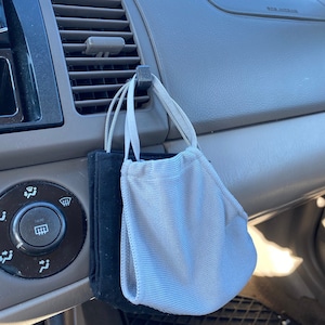 May include: Two face masks, one black and one light gray, hanging from a car vent. The masks are made of a soft, breathable material. The car's interior is a neutral color.