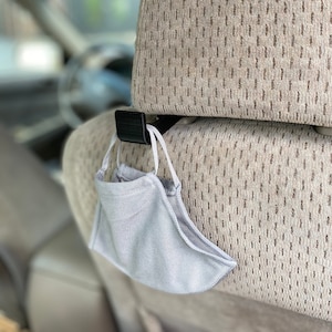 May include: A black plastic car headrest hook holds a light gray face mask with white ear loops. The mask is made of a soft, ribbed fabric. The headrest is beige with a dotted pattern.