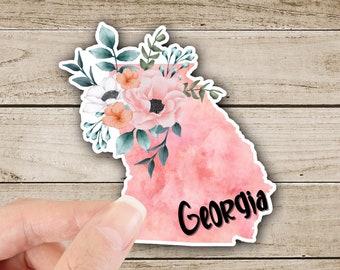 May include: A pink watercolour outline of the state of Georgia with a floral bouquet in the top left corner. The word "Georgia" is written in black lettering at the bottom of the outline.
