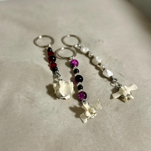 May include: Three handmade keychains with silver-tone rings. Each keychain features a small, off-white bone charm and a strand of beads in various colors: red and black, purple and black, and white. The keychains are displayed on a neutral-toned surface.