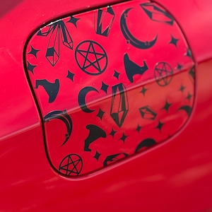 May include: A red car fuel door with a black pattern of crystals, moons, stars, and pentagrams. The design is repeated across the door's surface, creating a mystical and eye-catching aesthetic. The door is part of a red car.