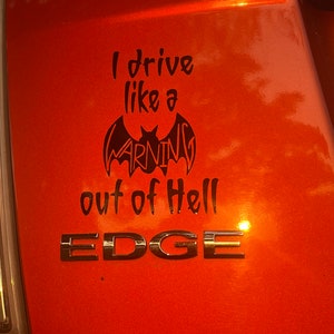 May include: Orange vehicle panel featuring a black decal with the text "I drive like a WARNING out of Hell." The word "WARNING" is within a bat shape. Chrome lettering spells out "EDGE" below the decal.