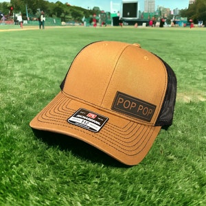 Könnte beinhalten: Eine braune und schwarze Baseballkappe mit schwarzem Netzrücken. Die Vorderseite der Kappe hat einen braunen Leder-Patch mit dem Text "POP POP" in weißen Buchstaben. Die Kappe ist von Richardson und hat einen verstellbaren Riemen.
