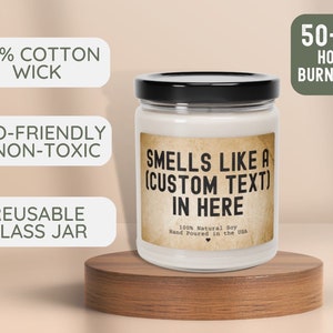 May include: A white candle jar with a black lid and a brown label that reads "Smells Like A (Custom Text) In Here". The label also says "100% Natural Soy Hand Poured in the USA". The jar is sitting on a wooden surface. The candle is eco-friendly, non-toxic, and has a 100% cotton wick. The jar is reusable and has a burn time of 50-60 hours.