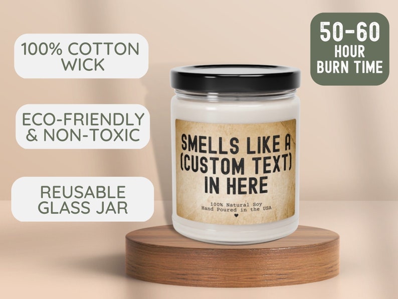 May include: A white candle jar with a black lid and a brown label that reads "Smells Like A (Custom Text) In Here". The label also says "100% Natural Soy Hand Poured in the USA". The jar is sitting on a wooden surface. The candle is eco-friendly, non-toxic, and has a 100% cotton wick. The jar is reusable and has a burn time of 50-60 hours.