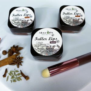 Może przedstawiać: Trzy małe słoiczki balsamu do ust z etykietą "Aksa Oils Fuller Lips" w czarnym kolorze z złotym akcentem. Słoiczki znajdują się na białym tle z brązowym pędzlem do makijażu i różnymi przyprawami. Smaki balsamu do ust to Sienna, Peach i Marron.