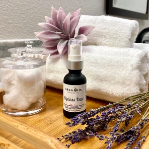 Może przedstawiać: Czarna butelka serum Akso Oils Ageless Dew z białą pompką, leżąca na drewnianej tacy z białymi ręcznikami, fioletowym kwiatem i suszoną lawendą.