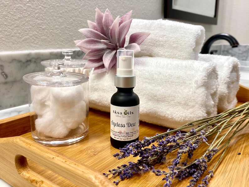 Może przedstawiać: Czarna butelka serum Akso Oils Ageless Dew z białą pompką, leżąca na drewnianej tacy z białymi ręcznikami, fioletowym kwiatem i suszoną lawendą.