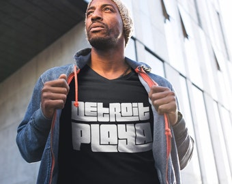 Könnte beinhalten: Eine Person trägt ein schwarzes T-Shirt mit dem Text "DETROIT PLAYA" in weißen Buchstaben. Die Person trägt eine Jeansjacke mit einem roten Reißverschluss und eine braune Mütze.