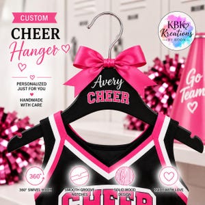 May include: A black wooden clothes hanger featuring a bright pink satin bow at the top. The hanger is personalised with the name Avery and the word CHEER in white and pink lettering. It includes a metal swivel hook and notched shoulders.