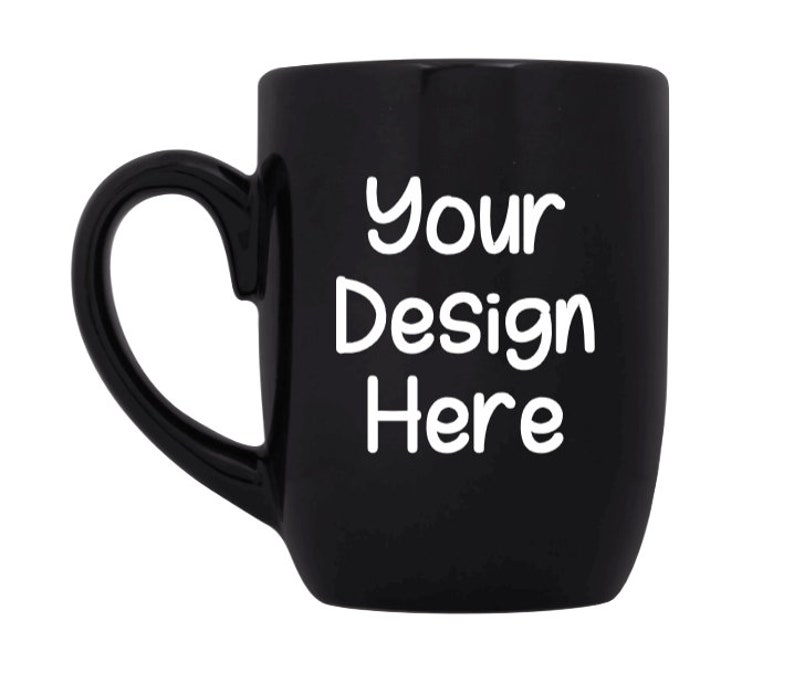 May include: A black ceramic mug with a white handle. The mug has the text "Your Design Here" printed on the front in a white, handwritten font.