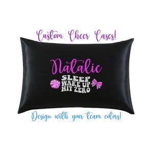 Op de afbeelding: Zwarte satijnen kussensloop met de naam "Natalie" in roze script, samen met de tekst "SLEEP WAKE UP HIT ZERO". De kussensloop heeft ook een roze pompon en strik. Tekst bovenaan zegt "Custom Cheer Cases!" en onderaan "Design with your team colors!"