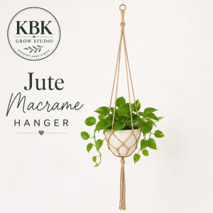 May include: A beige jute macrame plant hanger holding a white pot with a green pothos plant. The text KBK GROW STUDIO Jute Macrame HANGER is visible on the left. The hanger features a knotted net design with a long tassel at the bottom.