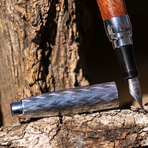 May include: A wooden and silver pen with a detailed design rests on a piece of weathered wood.