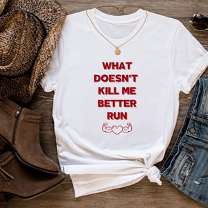 Puede incluir: Camiseta blanca con texto rojo que dice "What Doesn't Kill Me Better Run" con un corazón rojo y dos bíceps rojos a cada lado del corazón.