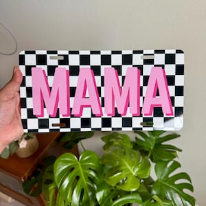 May include: A license plate with a black and white checkered background and the word "MAMA" in pink, outlined in a darker pink. The plate has three holes for mounting.