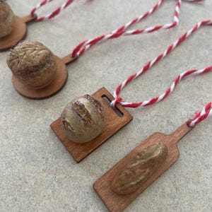 May include: Miniature bread ornaments, including a round loaf and a baguette, rest on small wooden cutting boards. Each ornament is attached to a red and white striped string, suggesting they are designed for hanging. The bread has a textured, golden-brown appearance.