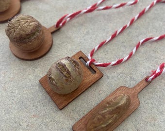 May include: Miniature bread ornaments, including a round loaf and a baguette, rest on small wooden cutting boards. Each ornament is attached to a red and white striped string, suggesting they are designed for hanging. The bread has a textured, golden-brown appearance.
