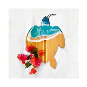 May include: A turquoise and white resin turtle-shaped cutting board with a wood base. The board is decorated with a wave design and a white sandy beach.
