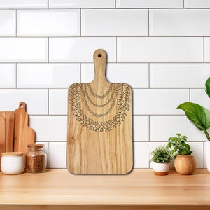 May include: A rectangular wooden cutting board with a handle. The board features a decorative design of layered necklaces and a leafy garland, etched in black. The board is made of light-colored wood and is displayed on a wooden countertop.