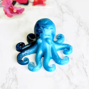 May include: A blue octopus ornament with a swirling design. The octopus has eight curled tentacles and a rounded head. The ornament is a gradient of light and dark blue. Pink flowers are in the background.