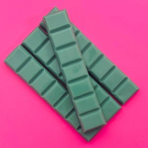 May include: Several teal-colored wax melt bars arranged diagonally against a vibrant pink background. Each bar is segmented into rectangular sections. The wax melts have a smooth, glossy appearance, suggesting a fresh, high-quality product.